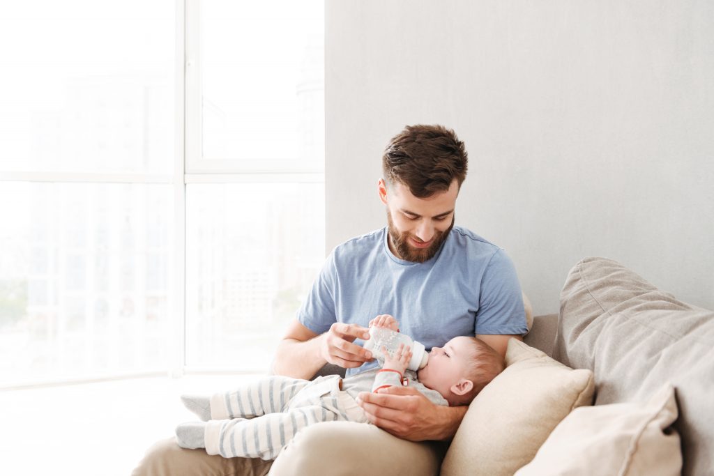 Image of young family. Father feeds her little child indoors at home.