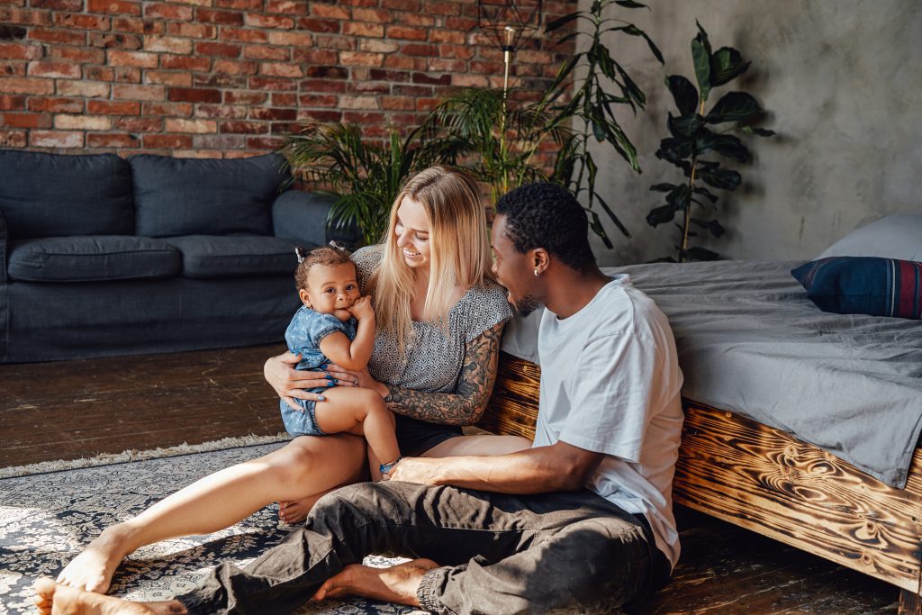 Shot of joyful parents with their little daughter sitting on floor inside living room in sunny day.