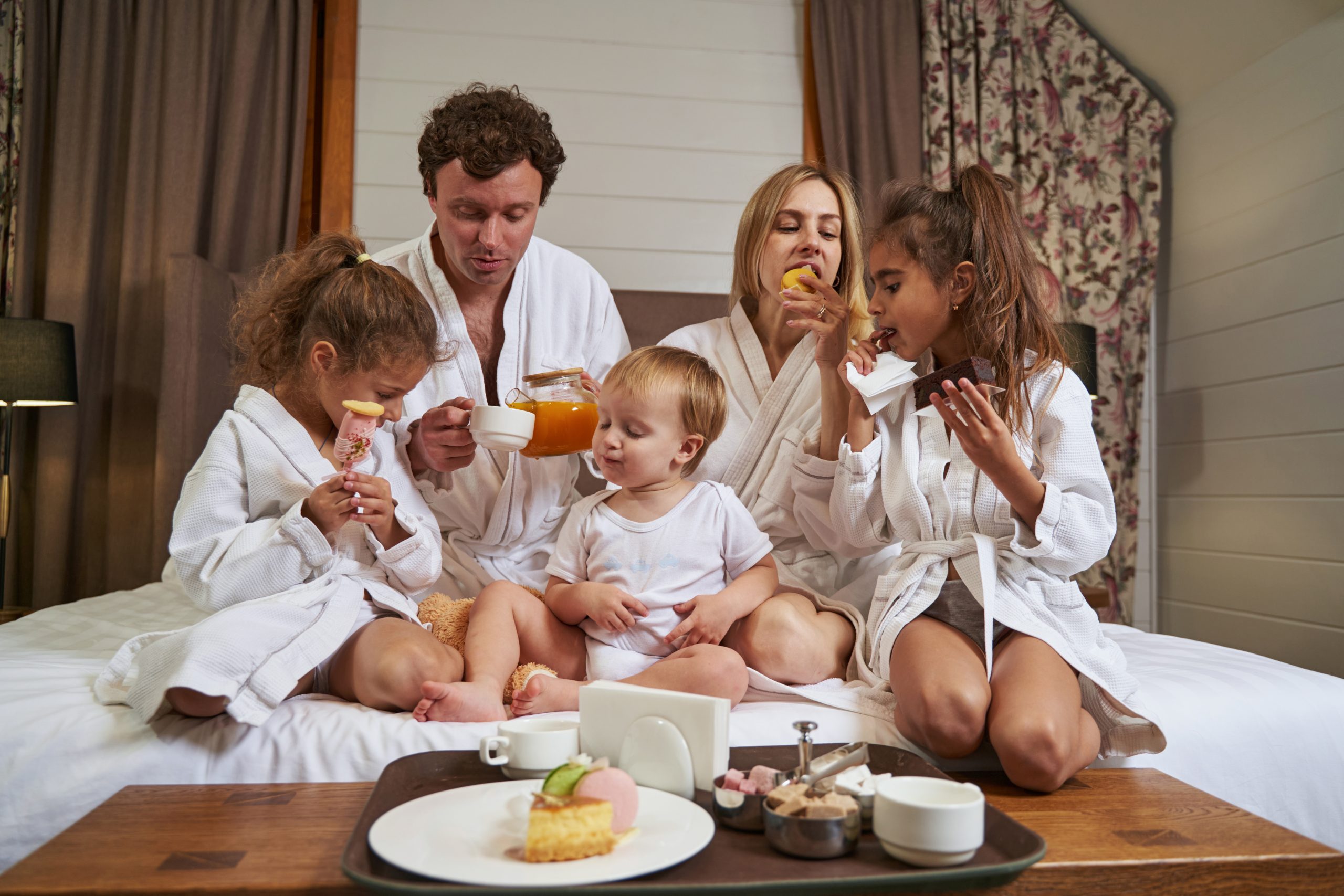 Young family in white coats sitting on bed near the table with food and having tasty breakfast